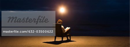 Man sitting in chair under light bulb on beach at night, reading book
