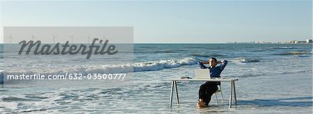 Man relaxing at desk set at water's edge on beach, wind turbines on horizon