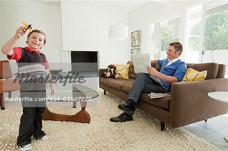 Young boy playing with toy aeroplane, father reading newspaper