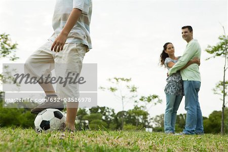 Boy playing with soccer ball, parents watching and embracing in background
