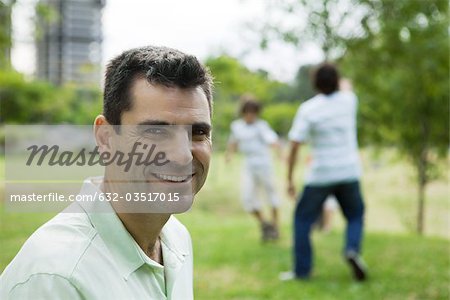 Man smiling at camera, sons playing in background