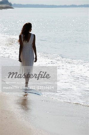 back view of woman wearing white dress and walking along the beach