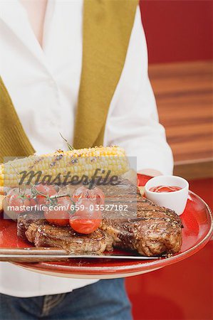 Woman holding T-bone steak, corn on the cob & cherry tomatoes