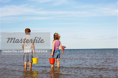 Brother and Sister in surf on beach with buckets and small fishnets looking out at ocean, back view