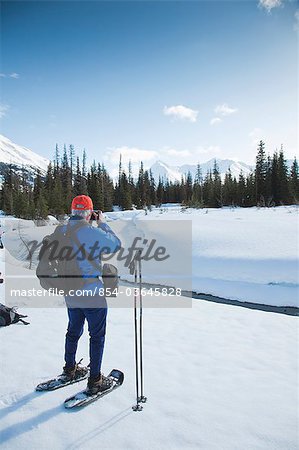 A mature man on snowshoes stops to enjoy the view beside Glacier Creek near the Johnson Pass Trail in the Kenai Mountain area of  Chugach National Forest, Kenai Peninsula, Southcentral Alaska, Spring