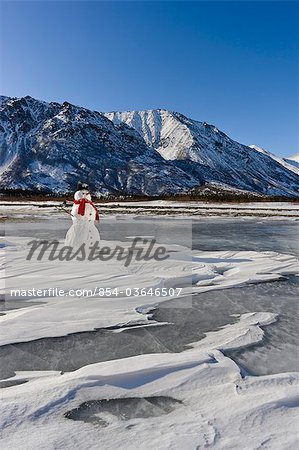 Schneemann mit einer roten Schal und schwarzen Hut sitzt auf dem gefrorenen Nenana River mit den Ausläufern der Alaskakette im Hintergrund, South Central Alaska, Winter