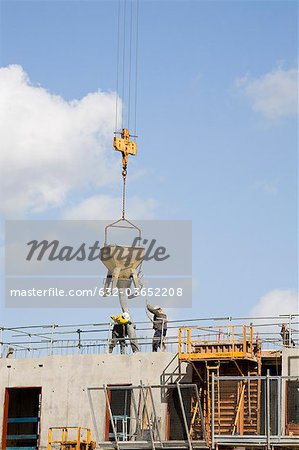 Construction workers guiding bucket suspended by crane to deposit concrete on building under construction