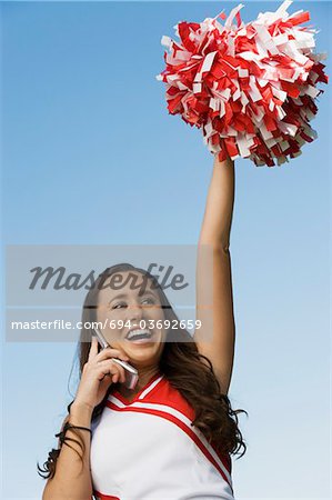 Smiling Cheerleader rising pom-pom, talking on mobile phone, (low angle view)
