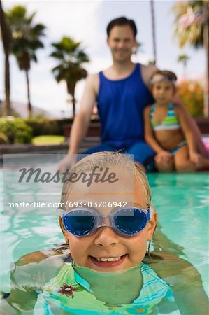 Girl (7-9) in swimming pool, father and sister (7-9) sitting on pool edge in background, front view portrait.
