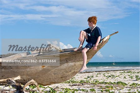 Kenya, Coast, Diani Beach.  A boy sits on a dug-out canoe whle on holiday on Galu Beach.