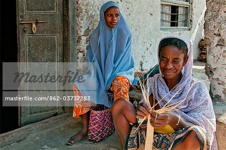 Tanzania, Zanzibar. Watched by her daughter, a Zanzibari woman weaves palm fronds to make baskets in Paje village.