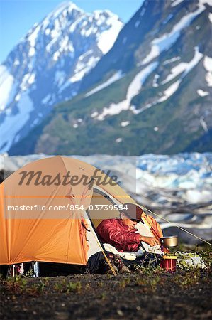 Femme prépare à manger sur un sac à dos poêle tandis que, dans sa tente avec Spencer Glacier et les montagnes Chugach à l'arrière-plan, forêt nationale de Chugach, centre-sud de l'Alaska, l'été