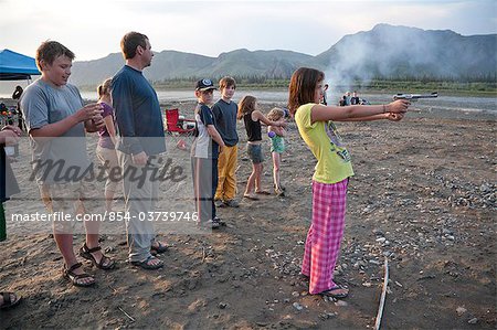 Ein 12 Jahres altes Mädchen steht und schießt eine.22 Pistole in ihrem Pyjama und Flip-flops am Rand des Yukon Rivers, während ein Floß hin-und Herbewegung Reise, Yukon-Charley Rivers National bewahren Interior Alaska, Sommer