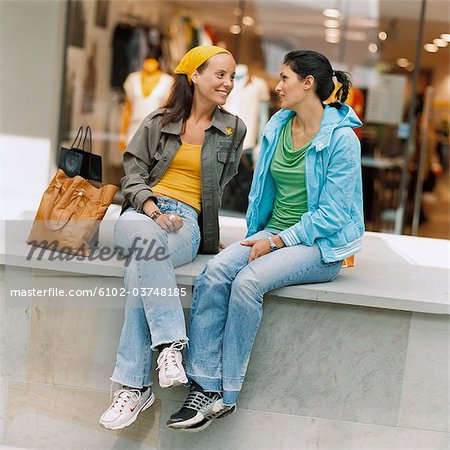 Two smiling women sitting in front of a shop-window.