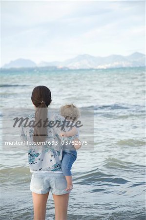 Une femme tenant un enfant en bas âge debout face à la mer avec les montagnes en arrière-plan lointain