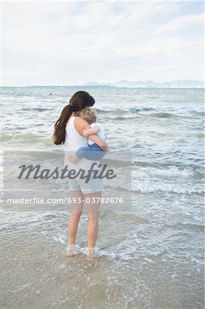 Une femme tenant un enfant en bas âge avec ses pieds dans l'eau peu profonde, debout face à la mer avec les montagnes en arrière-plan lointain