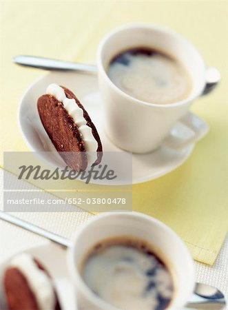 Chocolate and hazelnut shortbread biscuits filled with sugar ganache and cups of coffee Chocolate and hazelnut shortbread biscuits filled with sugar ganache and cups of coffee