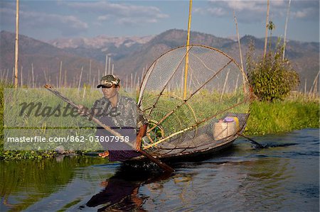 Myanmar, Inle-See. Intha Fischer mit traditionellen konischen Fische net, sanft sein Haus mit flachem Boden Boot paddeln.