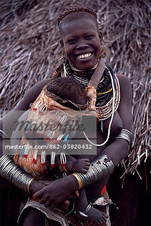A young Nyangatom woman carries her baby on her hip in an elaborately braided papoose. Her hair has been reddened with a mixture of ochre and animal fat. Typical of her tribe, she wears a calfskin skirt, multiple layers of bead necklaces and metal bracelets and amulets. The Nyangatom or Bume are a Nilotic tribe of semi nomadic pastoralists who live along the banks of the Omo River in south western