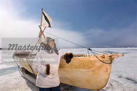 Mâle chasseur Inupiaq Eskimo, debout à côté d'un Inupiaq Umiaq fait de peau de phoque barbu (Ugruk) tout en portant une parka traditionnelle des Esquimaux (Atigi) et seal skin hat, mer des Tchouktches près de Barrow, Alaska arctique, été