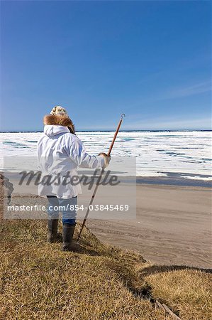 Portrait d'un chasseur Inupiaq Eskimo mâle portant son parka Eskimo (Atigi) et un chapeau de peau de phoque et tenant un bâton de marche à Utkeagvik ancien lotissement original avec vue sur la mer des Tchouktches, Barrow, Alaska arctique, l'été