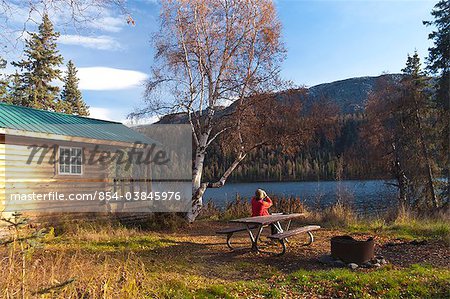 Frau genießt Blick auf Zugriffe durch Ferngläser während der Sitzung auf ein Picknick Tisch, Byers See öffentliche Nutzung Kabine, Denali State Park, South Central Alaska, Herbst