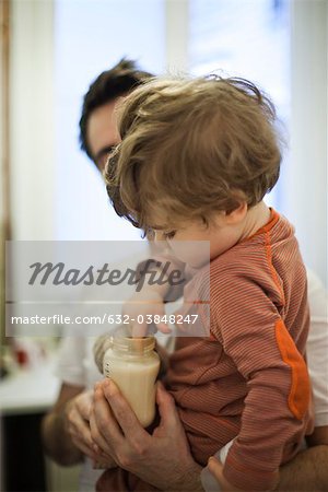 Toddler boy in father's arms, testing temperature of milk in baby bottle