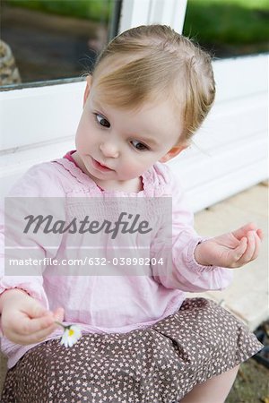 Toddler girl holding flower, looking down curiously, portrait