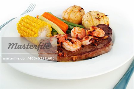 Meal setting of a steak with garlic butter sauce and fresh king prawns - surf n' turf - and fresh vegetables, on a white plate with knife and fork isolated on white