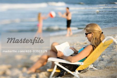 Caucasian mother reading in lounge chair on beach while husband and daughter play ball in background.