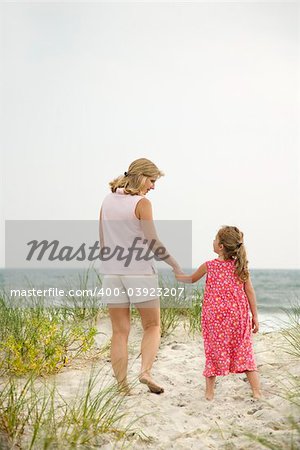 Caucasian mid-adult woman walking and holding hands with Caucasian female child on beach.