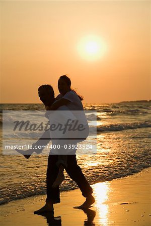 Caucasian mid-adult male carrying female piggyback on beach at sunset.