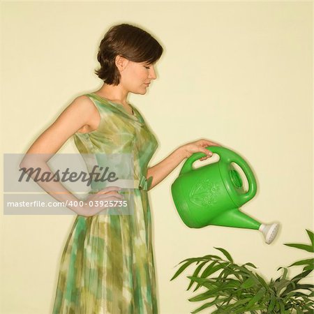 Pretty Caucasian mid-adult woman wearing vintage dress watering houseplant with green watering can.