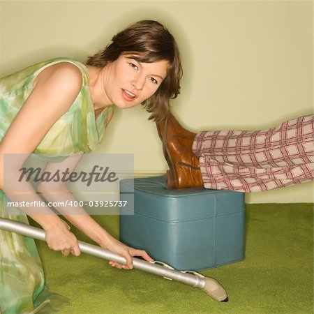 Unhappy pretty Caucasian mid-adult woman kneeling and vaccuuming carpet around male feet resting on foot stool.