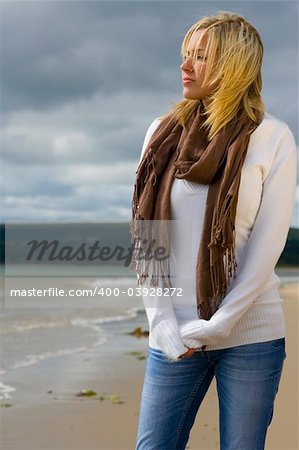 A beautiful blond haired blue eyed young woman standing alone on a beach deep in thought lit by golden sunlight but with stormy skies behind her