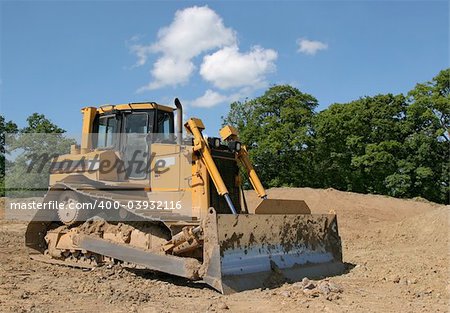 Yellow bulldozer standing idle on rough earth with trees and a blue sky to the rear.