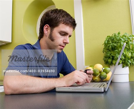 A young male shops online with a credit card at home in the kitchen.