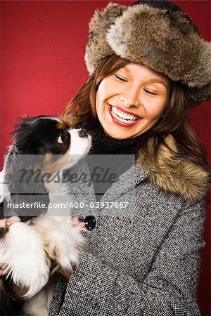 Young adult Caucasian woman wearing fur hat holding King Charles Spaniel in arms and smiling.