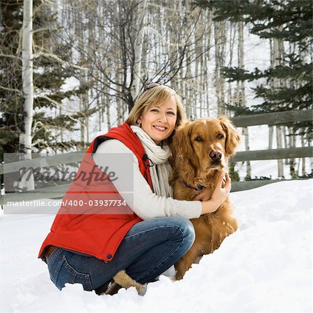 Attractive smiling mid adult Caucasian blond woman squatting in snow with arms around Golden Retriever.