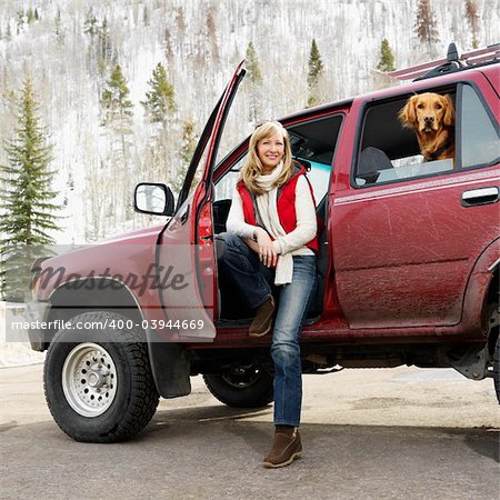 Woman sitting in dirt splattered SUV with door open as dog in back seat looks out open window in snowy countryside.