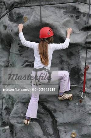 Young girl climbing upwards on a rock training face wearing a safety harness and red hard hat.