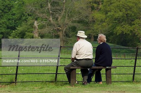 Elderly couple sitting together on a bench in front of a fence, gazing at a rural view .