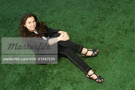 Young woman in the black jacket sitting cross legged on the green grass in the park, smiling