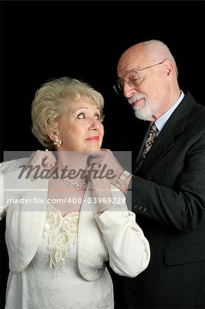 A handsome senior couple getting ready for a special occasion.  He is fastening her necklace for her.