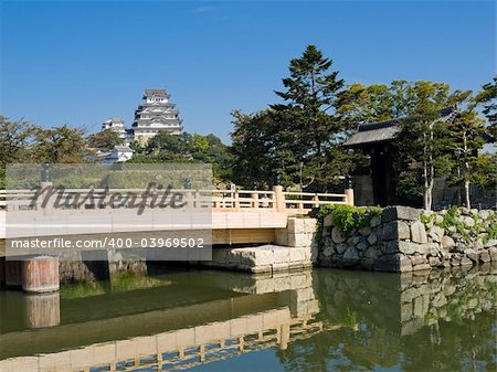Entrance to Himeji Castle grounds surrounded by a moat with main gate and bridge crossing in the foreground and Himeji Castle with clear blue sky in the background