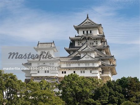 Landscape view of the main tower of Himeji Castle on the hillside during the daytime with trees of the castle grounds in the foreground and blue sky with thin white clouds in the background