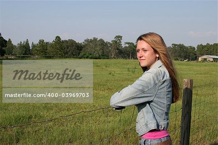 a beautiful girl on a ranch, leaning on a fence looking at a field, with a barn in the distance