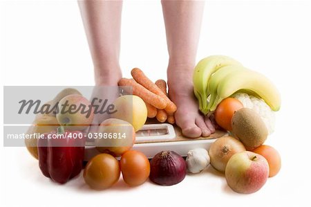 A pair of female legs standing on a scale surrounded by fruits and vegetables.