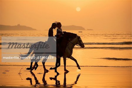 two people on horse back riding on wet sandy beach at sunset