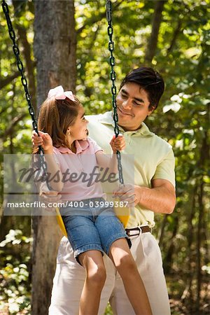 Hispanic father pushing daughter on swing and making eye contact.